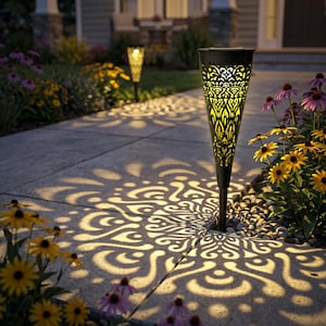 May include: Black solar-powered garden torch with intricate cut-out patterns, casting decorative light patterns on a concrete walkway. The torch is surrounded by yellow and purple flowers, creating a warm, inviting outdoor scene.