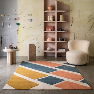 May include: A contemporary rug with a geometric design in yellow, blue, and orange. The rug has a cream background with diamond and rectangular shapes. The rug is in a room with a cream chair and a pink bookshelf. The room has a modern aesthetic.