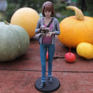 May include: A figurine of a person holding a vintage camera, wearing a gray jacket, pink shirt, and blue jeans. The figurine stands on a black base, with pumpkins and fruit in the background.