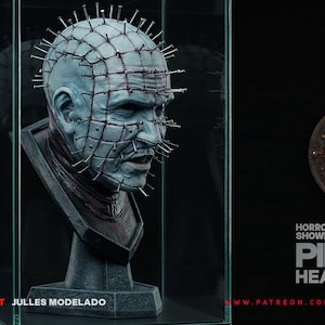 Könnte beinhalten: Detaillierte Büste von Pinhead aus der Hellraiser-Reihe, mit blassblauer Haut, zahlreichen Stiften im Kopf und einem düsteren Gesichtsausdruck. Die Skulptur wird in einer Glasvitrine mit dem Text "PINHEAD HEAD 1:1 SCALE" ausgestellt.