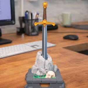 Op de afbeelding: Decoratief zwaardbeeldje met een gouden gevest en een blauw lemmet, geplaatst op een grijze stenen basis met een schedel. Het zwaard is ongeveer 20 cm hoog, een uniek bureauaccessoire.