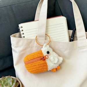 Puede incluir: Un bolso tote color crema con un pequeño monedero con forma de pato tejido a ganchillo en naranja y blanco adjunto. El monedero tiene una cremallera y un anillo dorado. Dentro de la bolsa hay un cuaderno y un bolígrafo. Una pequeña planta suculenta en una maceta blanca está en el fondo.