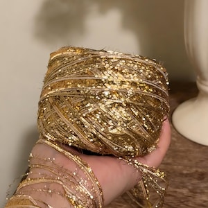 May include: A spool of gold sequin ribbon. The ribbon is tightly wound, with a shimmering, textured surface. The loose end of the ribbon is visible, suggesting it is ready for crafting or embellishment. The ribbon is a warm gold colour.