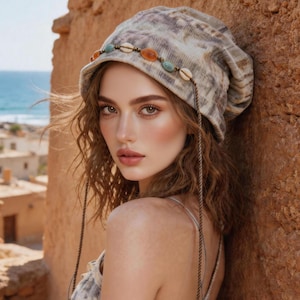 May include: A beige and gray patterned hat with a decorative band of shells and beads. The hat has long, dangling cords. The person in the image is wearing a matching patterned top. The background includes a stone wall and a view of the ocean.