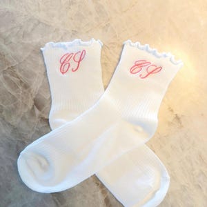 May include: A pair of white socks with a ribbed design. Each sock features a ruffled top and embroidered pink initials. The socks are displayed on a marble surface.