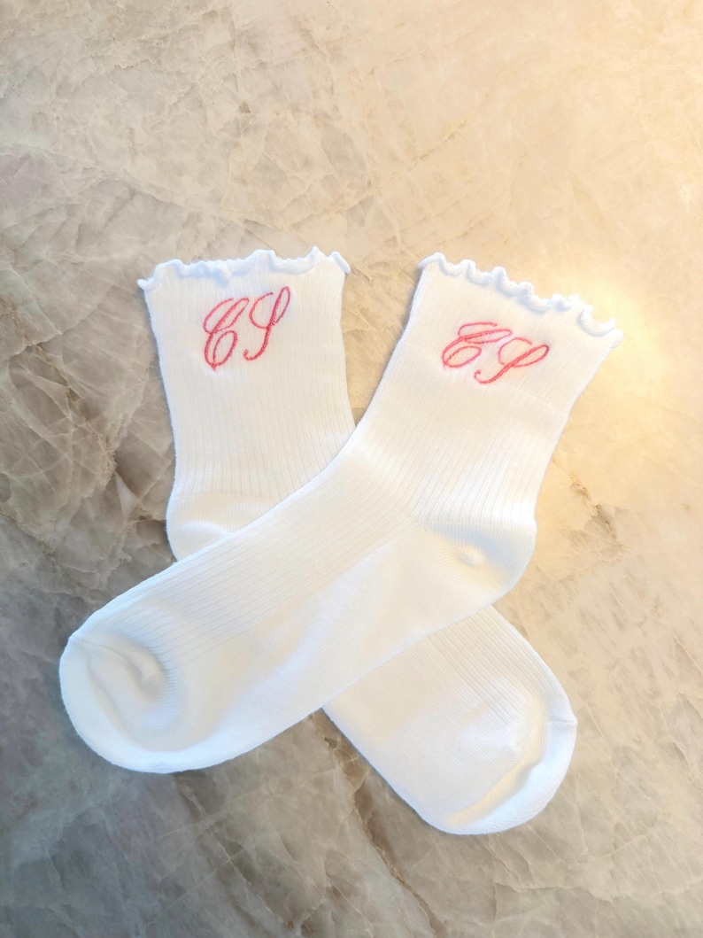 May include: A pair of white socks with a ribbed design. Each sock features a ruffled top and embroidered pink initials. The socks are displayed on a marble surface.