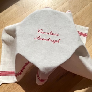 May include: White linen bread cloth with red stripes and the embroidered text "Caroline's Sourdough" in red script. The cloth is draped over a loaf of bread, with the corner of a second cloth visible.