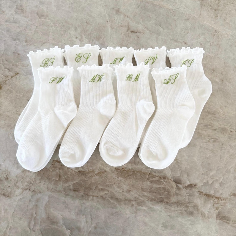 May include: A collection of white ankle socks with a ruffled top edge. Each sock is embroidered with a single initial in green thread. The socks are arranged on a light-colored surface.