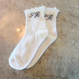 May include: A pair of white ribbed socks with ruffled tops. The socks have the embroidered letters "IR R" in black on the front. The socks are made of a soft, comfortable material.