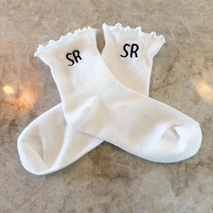 May include: A pair of white socks with a ruffled top edge. The socks have the embroidered letters "SR" in black near the top. The socks are folded over each other on a marble surface.