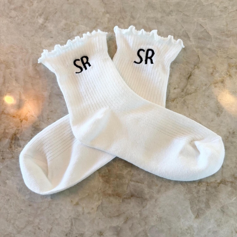 May include: A pair of white socks with a ruffled top edge. The socks have the embroidered letters "SR" in black near the top. The socks are folded over each other on a marble surface.