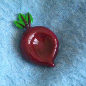 May include: A glossy, burgundy-colored beet-shaped brooch with green and red accents. The brooch is set against a light blue, textured background, creating a striking contrast. The design is a playful take on a vegetable.