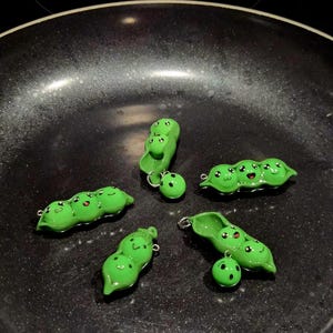 Peut inclure: Plusieurs breloques en forme de gousses de pois vertes avec des visages souriants et des détails rouges. Chaque gousse a une boucle argentée pour l'attacher à un bijou. Les pois sont disposés sur une surface sombre.