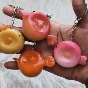 Op de afbeelding: Vier kleurrijke sleutelhangers in de vorm van een vis in geel, oranje, rood en roze. Elke sleutelhanger heeft een zilveren ketting en ring. De vissen hebben kleine vinnen en een rond lichaam. De sleutelhangers worden in een hand gehouden.