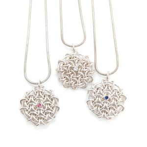 May include: Three silver necklaces with woven circular pendants. Each pendant features a central gemstone: a clear stone, a pink stone, and a blue stone. The necklaces hang against a white background.
