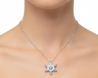 Op de afbeelding: Een zilveren ketting met een sneeuwvlokhanger. De hanger is versierd met lichtblauwe en heldere edelstenen, gerangschikt in een symmetrisch patroon. De ketting wordt getoond tegen een neutrale achtergrond, waardoor het ontwerp van de sieraden wordt benadrukt.