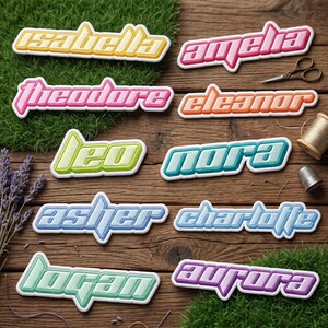 May include: A collection of ten custom name patches featuring various colors and fonts, including Isabella, Amelia, Theodore, Eleanor, Leo, Nora, Asher, Charlotte, Logan, and Aurora, arranged on a wooden surface with scissors and thread.