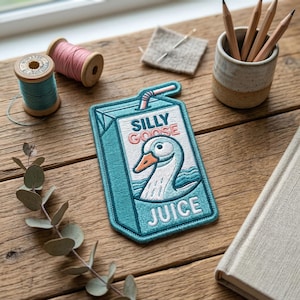 May include: A teal embroidered patch in the shape of a juice box, featuring a cartoon goose and the words "SILLY GOOSE JUICE". The patch has a pink straw and text. Other items on a wooden surface include thread spools, pencils, and a small ceramic cup.