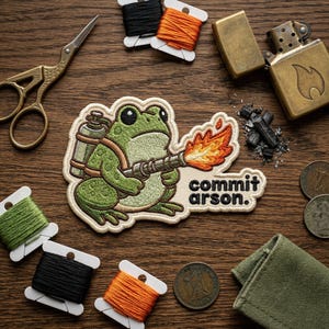 May include: Embroidered patch featuring a green frog with a flamethrower, the text "commit arson." is below the frog. The patch is surrounded by spools of thread, scissors, a lighter, and coins on a wooden surface.