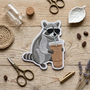 May include: Embroidered patch featuring a raccoon wearing sunglasses and holding a coffee cup with a straw. The raccoon is gray and white with black sunglasses. The coffee cup is brown. The patch is on a wooden surface.
