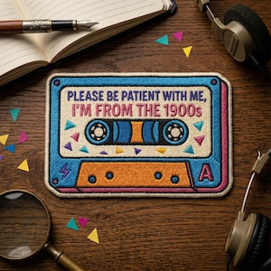 May include: Embroidered patch in the shape of a cassette tape with the text "PLEASE BE PATIENT WITH ME, I'M FROM THE 1900s". The patch has a blue border, orange center, and colorful accents. A retro-themed accessory.