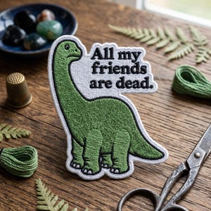 May include: Embroidered patch featuring a green dinosaur with the text "All my friends are dead." The dinosaur is outlined in white, with a detailed stitched texture. The patch is on a wooden surface.
