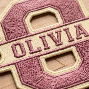 May include: A custom embroidered patch featuring the letter O in a dusty rose color with a cream-colored border. A horizontal cream-colored banner runs across the center of the letter, displaying the name OLIVIA in dusty rose embroidered letters.