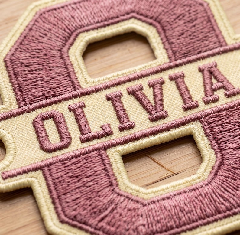 May include: A custom embroidered patch featuring the letter O in a dusty rose color with a cream-colored border. A horizontal cream-colored banner runs across the center of the letter, displaying the name OLIVIA in dusty rose embroidered letters.