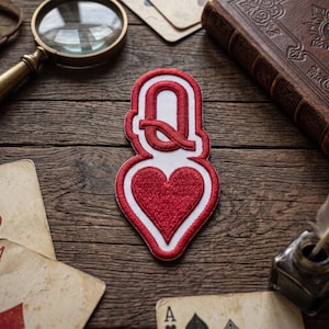 May include: A red and white embroidered patch featuring the letter Q positioned above a heart shape, designed to resemble a playing card symbol. The patch has a thick red border and a white background, with the letter Q and heart in red thread.