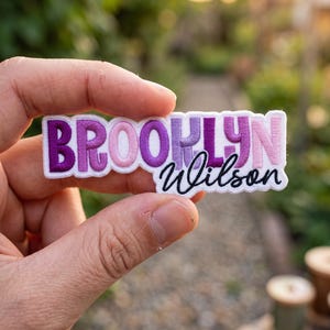 May include: A rectangular embroidered patch featuring the name BROOKLYN in a bold, sans-serif font with a gradient of purple and pink thread colours, followed by the name Wilson in a black cursive script font on a white fabric background.