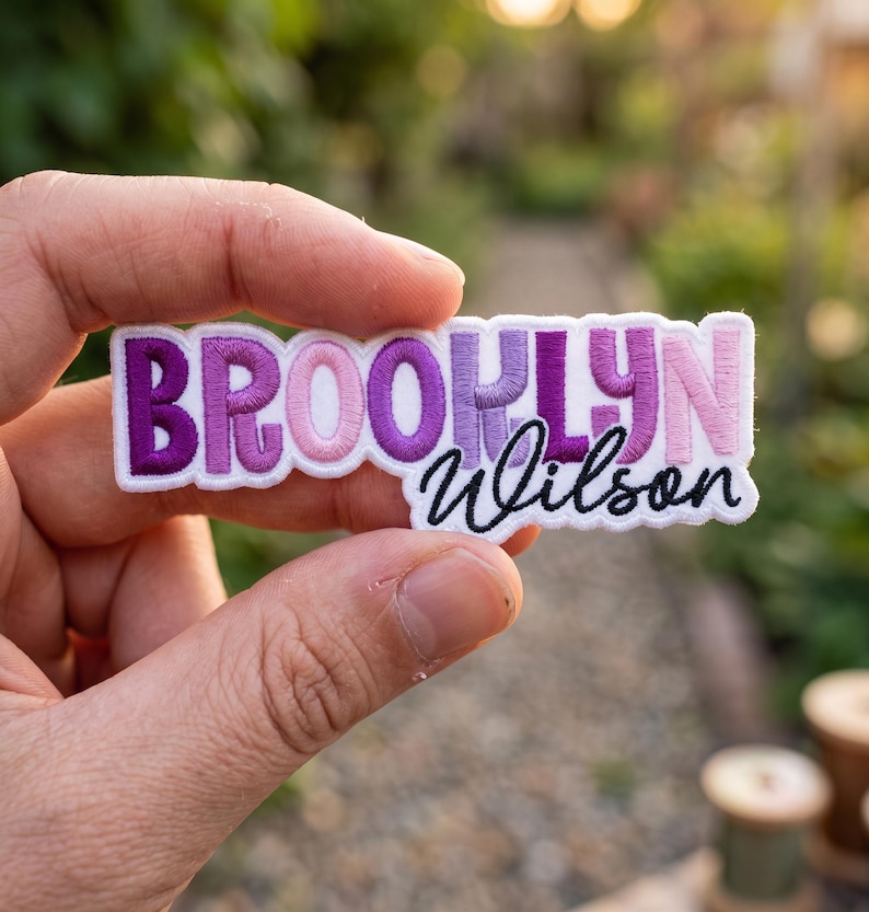May include: A rectangular embroidered patch featuring the name BROOKLYN in a bold, sans-serif font with a gradient of purple and pink thread colours, followed by the name Wilson in a black cursive script font on a white fabric background.
