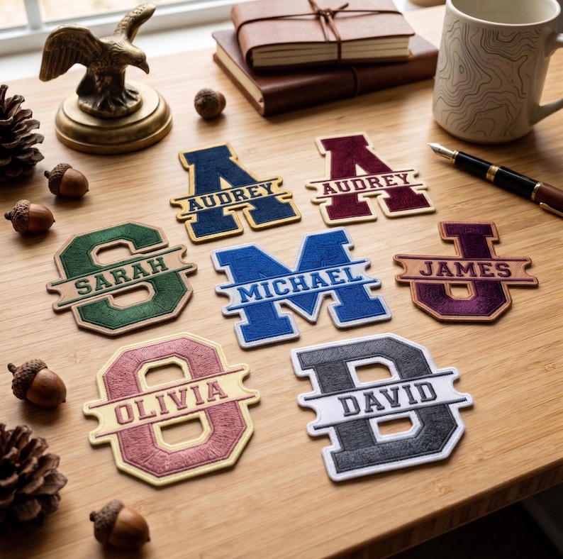 May include: A collection of seven embroidered fabric patches, each featuring a large block letter with a horizontal banner displaying a name. The names include Audrey, Sarah, Michael, James, Olivia, and David, rendered in various colors.