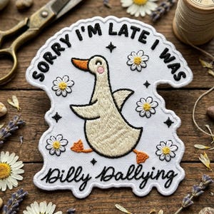 May include: Embroidered patch with a white background and a cartoon goose design. The patch features the text "SORRY I'M LATE I WAS" and "Dilly Dallying" in black script. Daisies and stars surround the goose.