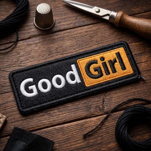 May include: A rectangular embroidered patch with a black border and background. The word Good is stitched in white thread, while the word Girl is stitched in black thread against a gold-colored rectangular background within the patch.