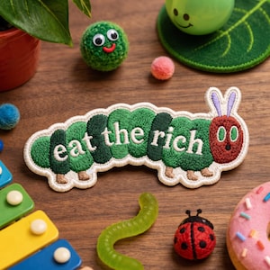 May include: An embroidered patch shaped like a green caterpillar with a red head and purple antennae. The body segments feature the white embroidered text "eat the rich" in a rounded font. The patch has a white border and brown feet.