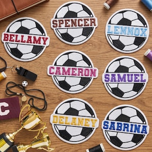 May include: Embroidered soccer ball patches with names: Bellamy, Spencer, Lennox, Cameron, Samuel, Delaney, and Sabrina. Each patch features a black and white soccer ball design with a name in a contrasting color. A whistle, trophy, and other soccer-related items are also visible.