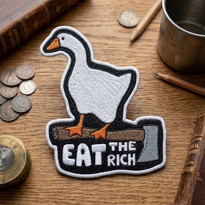May include: Embroidered patch featuring a white goose with orange feet and beak, standing on a log with an axe. The patch has the text "EAT THE RICH" in white letters with a black border. The patch is on a wooden surface.