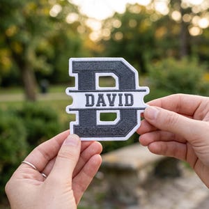 May include: A dark gray and white embroidered patch in the shape of the letter D, featuring a horizontal white bar across the center with the name DAVID stitched in dark gray capital letters, surrounded by a thick white border.