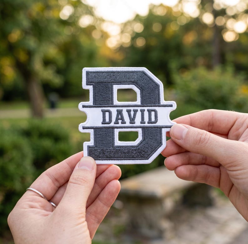 May include: A dark gray and white embroidered patch in the shape of the letter D, featuring a horizontal white bar across the center with the name DAVID stitched in dark gray capital letters, surrounded by a thick white border.
