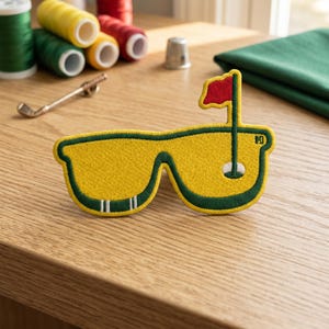 May include: Embroidered patch in the shape of sunglasses with a golf flag design. The patch features a yellow lens with a green border and a red flag. The design includes a golf hole and the letter "H".