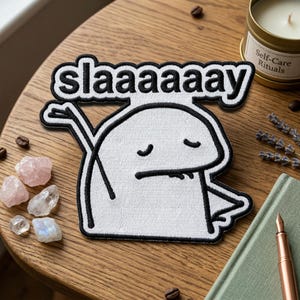 May include: A white patch with a black outline featuring a cartoon character with closed eyes and the word "slaaaaaay" in black. The patch is on a wooden surface with crystals, a candle, and a pen.