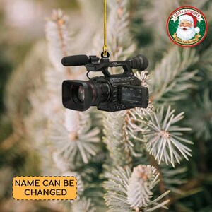 May include: A black video camera Christmas ornament hanging from a gold string. The ornament has a microphone and a screen. A Santa Approved sticker is in the upper right corner. The text "NAME CAN BE CHANGED" is on a yellow rectangle.