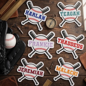 May include: Baseball-themed patches with names embroidered in various colors. Each patch features a baseball design with crossed bats and a name: Harlan, Teagan, Frankie, Thaddeus, Jeremiah, and Malachi. The patches are on a wooden surface.