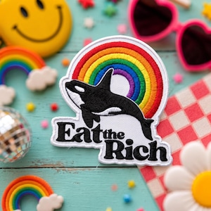 May include: A white embroidered patch featuring a black and white orca whale leaping in front of a vibrant, multi-colored rainbow arch. Below the orca, the text Eat the Rich is stitched in bold, black, capitalized letters on a white background.
