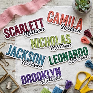 May include: A collection of personalized name stickers in various colors, including red, orange, green, blue, and purple. Each sticker features a name and the last name "Wilson" in a decorative font. The stickers are arranged on a white fabric surface.