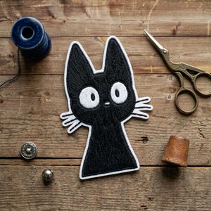 May include: Black embroidered cat patch with white details, featuring large, round eyes and whiskers. The patch is on a wooden surface, alongside sewing tools like scissors, a thimble, and thread.
