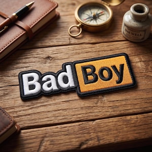 May include: A rectangular embroidered patch featuring the text Bad Boy. The word Bad is in white letters on a black background, and the word Boy is in black letters on a mustard yellow background. The entire patch has a thick black border.
