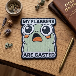 May include: Embroidered patch featuring a cartoon frog with large eyes and an open mouth, expressing surprise. The text "MY FLABBERS ARE GASTED" is displayed above and below the frog. The patch is green, white, black, and orange.