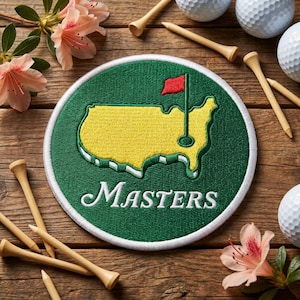 May include: A round green and white embroidered patch with the word "Masters" in white script. The patch features a yellow map of the United States with a red flag on a golf course. Wooden golf tees, golf balls, and pink flowers are scattered around the patch.