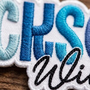 May include: An embroidered patch featuring the letters CHS in varying shades of blue thread on a white background. Below the blue letters, the word uli is stitched in black cursive thread. The patch has a white scalloped border edge.
